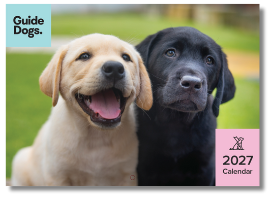 Image of a 2027 wall calendar cover featuring two Labrador retriever puppies outdoors on a grassy background, with the Guide Dogs logo in the top-left and a pink '2027 Calendar' label in the bottom-right.