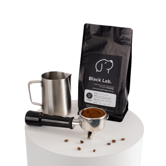 Image of A bag of Soho Coffee and Hot Chocolate with a metal milk pitcher and portafilter on a white round table.