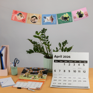 A desk with calendars featuring dogs, a plant in a green pot, office supplies, and cards with dog photos hanging on the wall. The desk calendar shows April 2026.