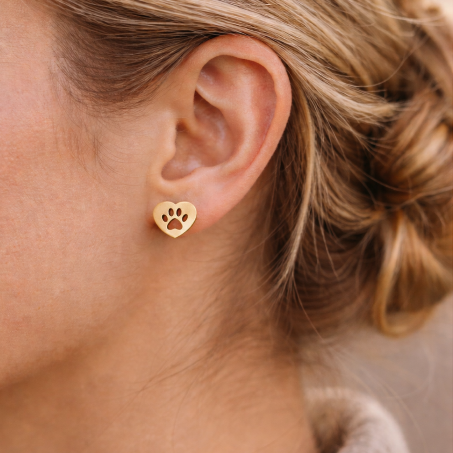 Image of a close-up of a woman wearing gold, heart shaped earrings with a paw print cutout.