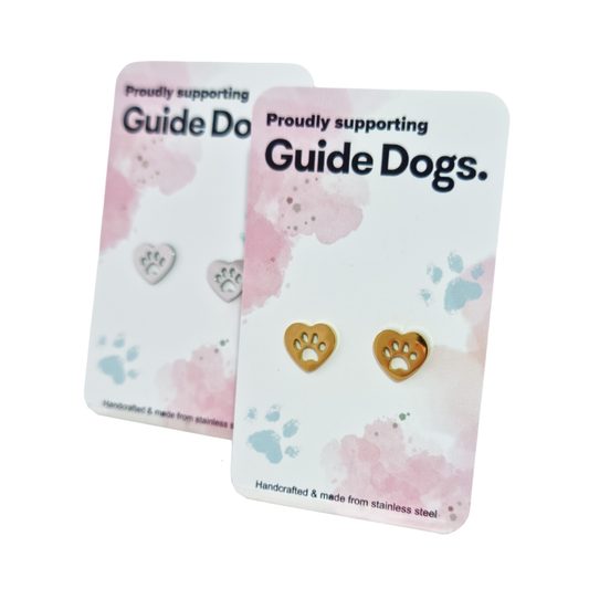 Image of a pair of gold, heart shaped earrings with a paw print cutout on pink and white packaging with Guide Dogs branding