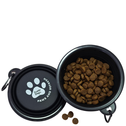 Image shows a black collapsible pet bowl shown open with kibble inside, alongside the matching lid. The lid has a white paw print and the text 'Guide Dogs' and 'PAWS AND REFRESH.