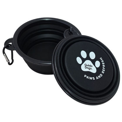 Image of a black collapsible pet water bowl with a collapsed bowl placed beside it on a white background. The collapsed bowl shows a white paw print logo with the text 'Guide Dogs' and 'PAWS AND REFRESH,' plus a small carabiner attached to the rim.