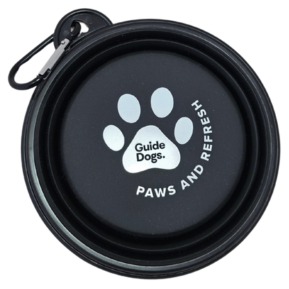 Image of a black collapsible dog water bowl with a carabiner clip, shown closed. The inside features a white paw print logo with the text 'Guide Dogs.