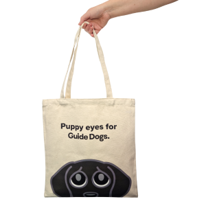 Image of a hand holding the Tote Bag - Puppy Eyes featuring a cartoon dog face and "Puppy eyes for Guide Dogs" text.