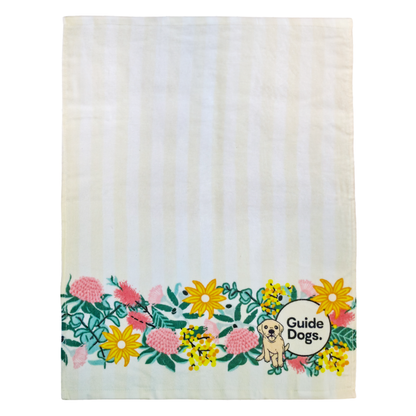 Image of a cream and white tea towel with  Guide Dogs logo and a dog illustration with floral elements on a white background.