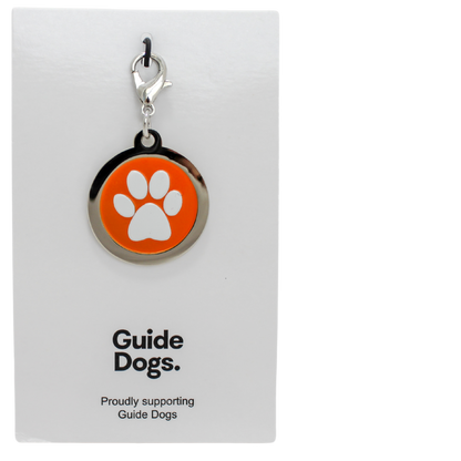 Image of a round metal charm with a white paw print on an orange background. on a card reading Guide Dogs.