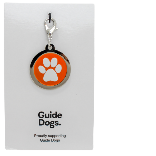 Image of a round metal charm with a white paw print on an orange background. on a card reading Guide Dogs.
