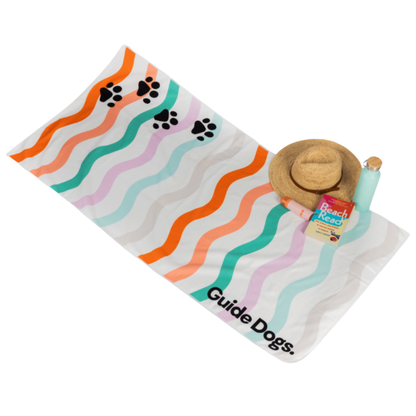 Image of beach towel with colorful wave pattern and 'Guide Dogs' text, featuring a sun hat and 'Beach Rose' product.