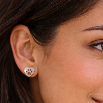 Image of a close-up of a woman wearing silver, heart shaped earrings with a paw print cutout.