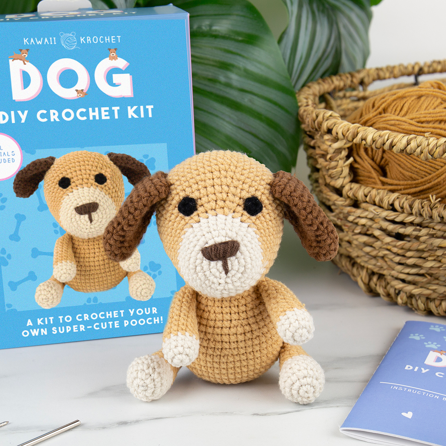Image of The Crochet Kit includes a crochet dog plush displayed before its DIY kit box, with matching yarn, basket, and instruction booklet on a white surface with green leaves—ideal for beginner crochet enthusiasts.