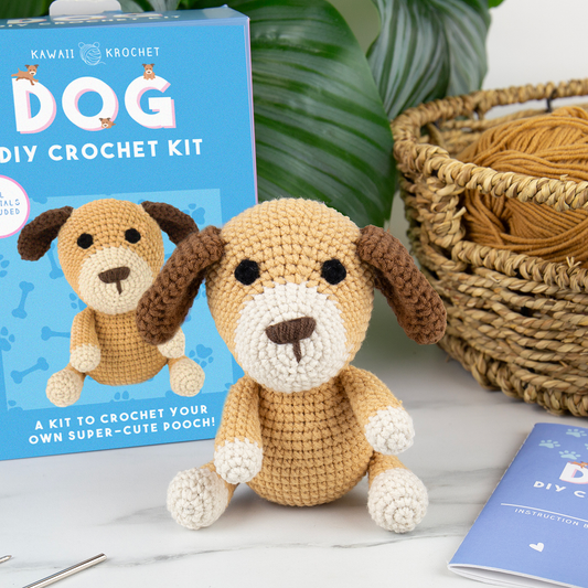 Image of The Crochet Kit includes a crochet dog plush displayed before its DIY kit box, with matching yarn, basket, and instruction booklet on a white surface with green leaves—ideal for beginner crochet enthusiasts.