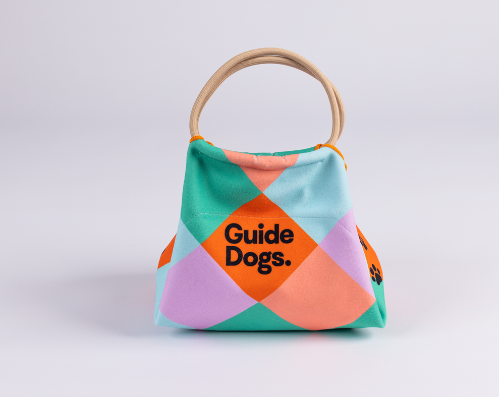 Image of a lunch bag with colourful squares, Guide Dogs text, and round tan handles on a light background.