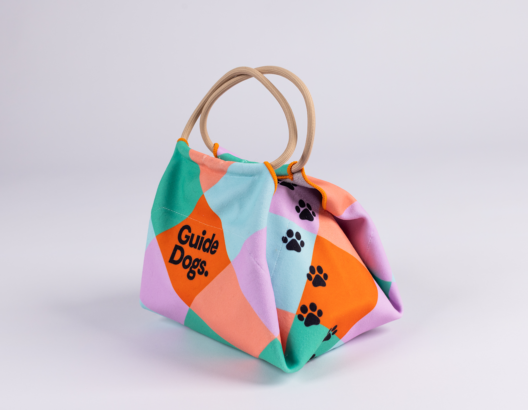 Image of a lunch bag with black paw prints up the side, "Guide Dogs" text, looped handles, and a colorful geometric pattern.