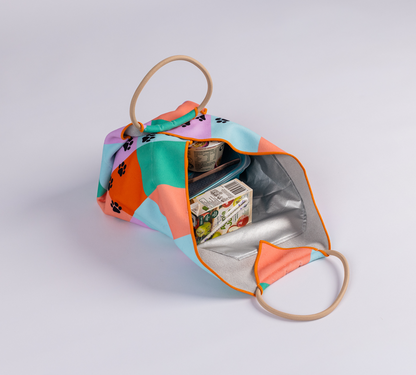 Image of a colourful reusable lunch bag with paw prints lies open, showing a small container of food and a juice box inside. The bag has round handles and an orange trim.
