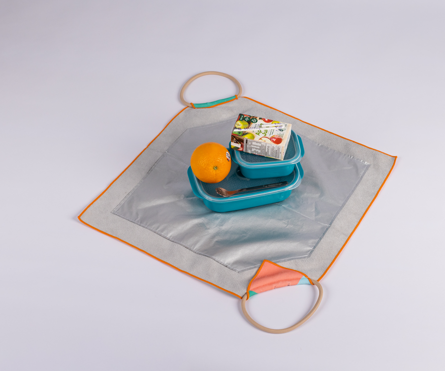 Image of a silver and orange square mat with handles at two corners displays a lunch set: a blue reusable lunch bag, an orange, and a packaged food item, all arranged neatly in the center.

