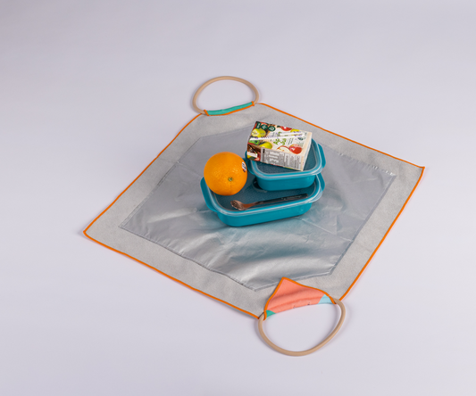 Image of a silver and orange square mat with handles at two corners displays a lunch set: a blue reusable lunch bag, an orange, and a packaged food item, all arranged neatly in the center.

