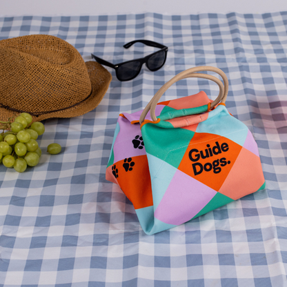 Image of a colourful reusable lunch bag with Guide Dogs written on it sits on a blue and white checkered picnic cloth, beside sunglasses, a straw hat, and a bunch of green grapes.