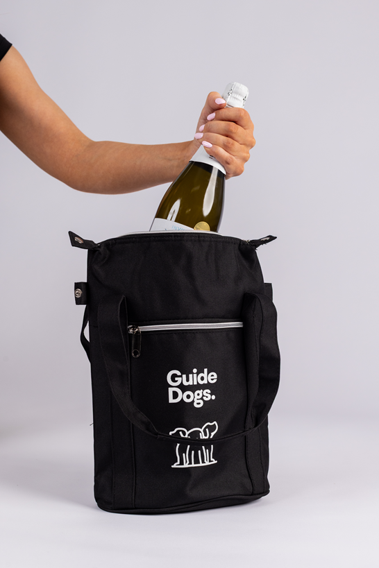 Image of a hand placing a wine bottle into a black Wine Cooler Bag, featuring Guide Dogs and a dog logo on the front.