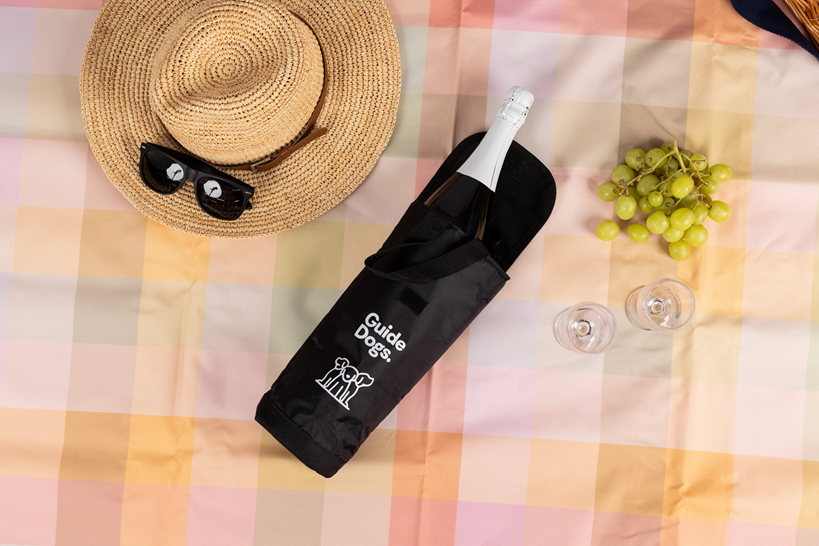 Image of a picnic setup on a plaid blanket features a black Wine Cooler Bag  labeled Guide Dogs holding a wine bottle.
