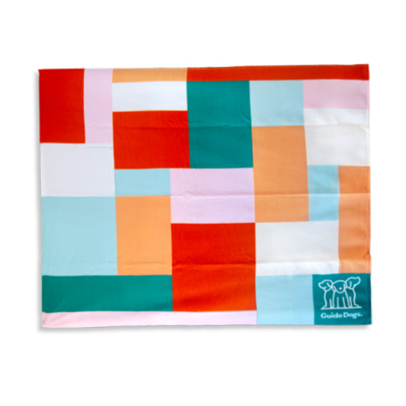 Image of a colorful geometric-patterned polar fleece blanket with a green corner featuring a white Guide Dogs logo.
