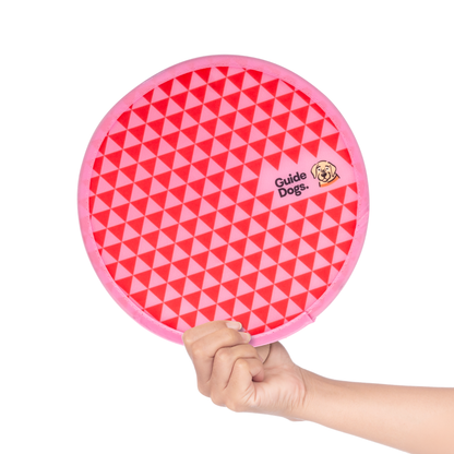Image of a hand holding a pink and red triangular-patterned Twist Fan labeled Guide Dogs with a dog illustration.